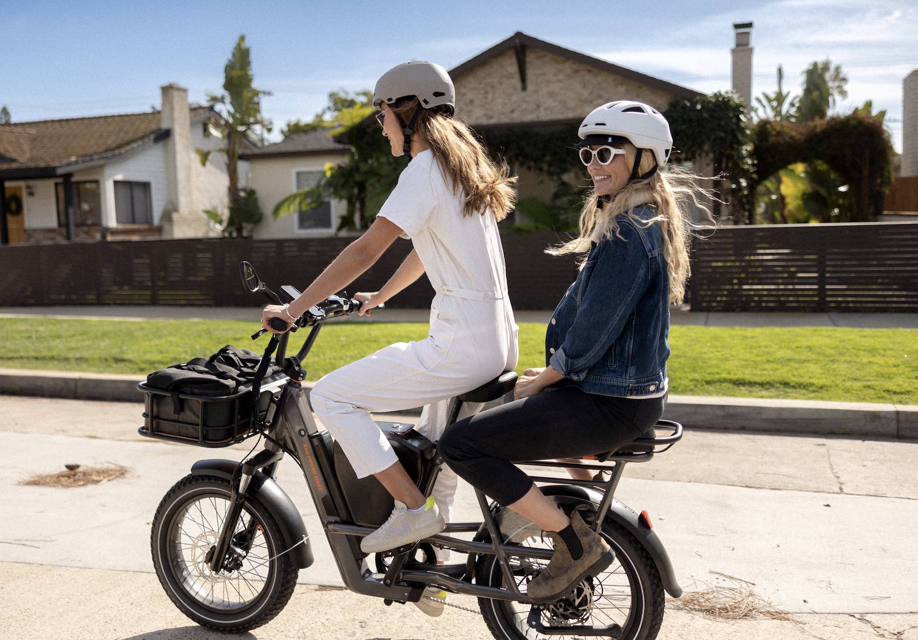 Two women ride a RadRunner 3 Plus on a suburban street in sunny weather.